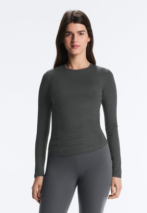 Donna in piedi, di fronte, indossa una maglia aderente a maniche lunghe grigio scuro e pantaloni abbinati su sfondo bianco uniforme.