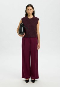 Gilet smanicato in maglia bordeaux con bottoni, abbinato a pantaloni larghi bordeaux. Tiene una borsa in pelle nera e si trova su uno sfondo neutro.
