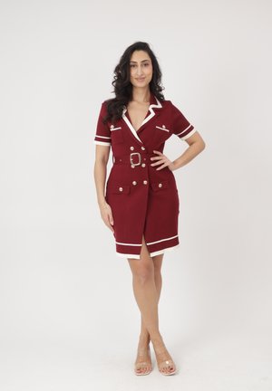 Mujer posando con confianza, vestida con un vestido burdeos con cinturón, ribete blanco y botones dorados, combinado con tacones de tiras transparentes.