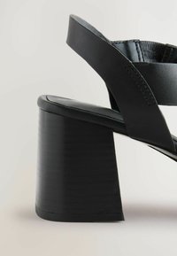 Mustat korkeat sandaalit, joissa on paksu pohja, sileä pinta ja ristikkäiset hihnat. Korko on neliön muotoinen ja puumaista pintakäsittelyä.