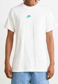 Camiseta de algodón blanca con cuello redondo y mangas cortas, que presenta un pequeño logo verde de Nike en el pecho. Ajuste ligeramente holgado.