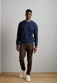 Navy blue cotton sweatshirt, brown trousers, and white sneakers. Model stands against a light gray wall on a wooden floor.