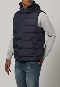 Mann trägt eine navyblaue gepolsterte Weste über einem hellgrauen Sweatshirt und blauen Jeans, mit einer Hand in der Tasche und dem Kopf leicht nach links gedreht.