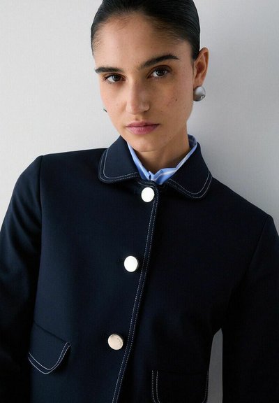 Femme aux cheveux tirés en arrière, portant de grandes boucles d'oreilles en perles et une veste bleu marine avec des surpiqûres blanches et trois boutons argentés, regardant la caméra.