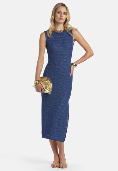 Femme portant une robe midi en maille bleue sans manches, un collier en chaîne dorée, un bracelet et une montre en or, tenant une pochette dorée, debout devant un fond blanc.