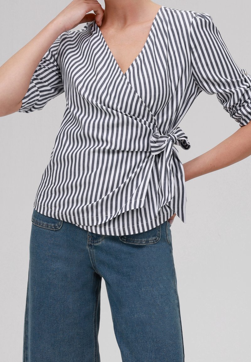 Blouse portefeuille rayée en noir et blanc avec un col en V, des manches bouffantes et une taille nouée. Associée à un jean à jambes larges bleu clair.