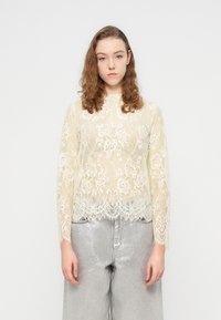 Haut à manches longues en dentelle couleur crème clair avec motifs floraux et ourlet festonné ; porté sur un pantalon large argenté, doté d'une texture lisse et brillante.