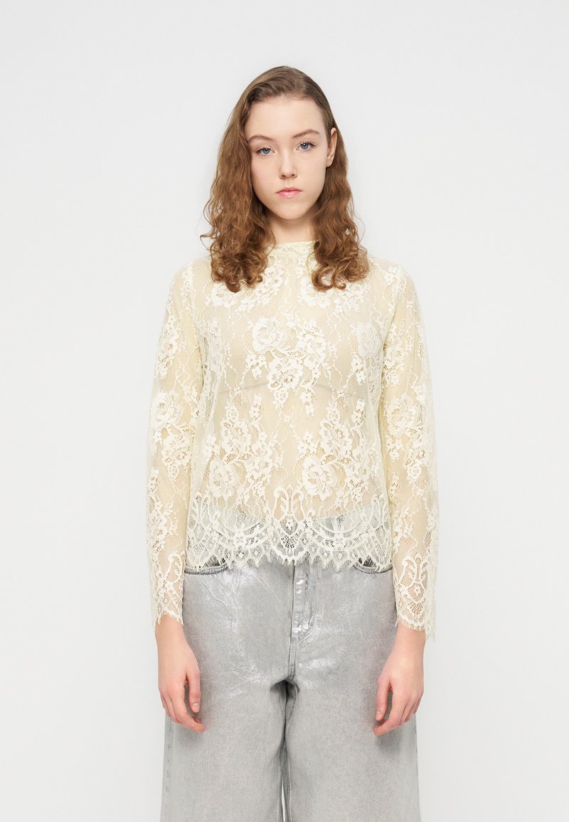 Haut à manches longues en dentelle couleur crème clair avec motifs floraux et ourlet festonné ; porté sur un pantalon large argenté, doté d'une texture lisse et brillante.