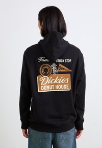 Hanorac negru cu un print grafic pe spate, având o ilustrație cu o gogoasă, "Dickies Donut House", și alte texte în bej și alb.