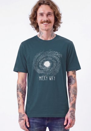Glimlachende man met schouderlang haar en tatoeages draagt een donker T-shirt met een witte Melkweg-galaxie afbeelding en tekst.