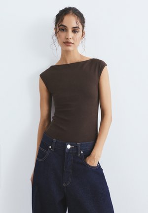 Kvinde med mørkt krøllet hår iført en tætsiddende mørkebrun ærmeløs top og løse mørkeblå jeans, den ene hånd i lommen, mod en ensfarvet baggrund.