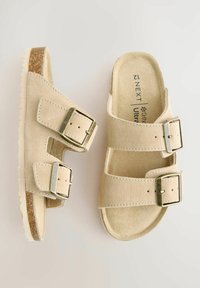 Beige Wildledersandalen mit zwei verstellbaren Riemen, silbernen Schnallen und einer korkähnlichen Sohle. Glatte Textur mit flachem, offenem Design.