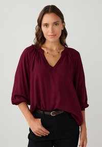Frau mit schulterlangem braunem Haar, die eine burgunderfarbene V-Ausschnitt-Bluse, schwarze Hose, einen braunen Gürtel und eine goldene Halskette trägt, vor einem schlichten Hintergrund stehend.