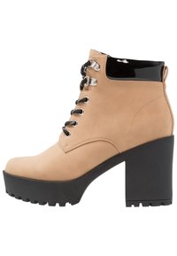 Botín de tobillo beige de ante con un panel negro brillante, diseño con cordones, herrajes plateados y un tacón de plataforma negro robusto.