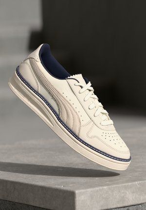 Tenis din piele crem, cu căptușeală de sosete albastră închisă, vârf perforat și cusături contrastante de-a lungul mijlocului și laterale.
