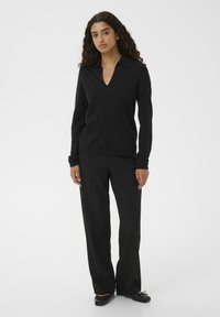Donna con lunghi capelli ricci che indossa una camicia nera a maniche lunghe con colletto, pantaloni neri a gamba larga e ballerine nere, in piedi davanti a uno sfondo bianco.