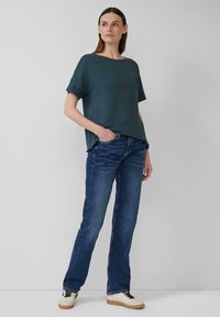 Donkerblauw groene kortemouwen top gecombineerd met blauwe jeans en beige sneakers. De top heeft een relaxte pasvorm en een ronde halslijn.