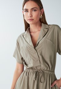 Giovane donna che indossa un vestito beige con bottoni e vita regolabile, con orecchini a cerchio dorati, in posa con una mano in tasca su uno sfondo semplice.