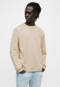 Μπεζ φούτερ με στρογγυλή λαιμόκοψη από βαμβακερό μείγμα ύφασμα, με ριμπ μανσέτες και τελείωμα, με ένα μικρό λογότυπο στο στήθος. Ορατό ανοιχτό μπλε τζιν.