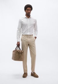 Camisa blanca abotonada combinada con pantalones beige. Sosteniendo una bolsa de viaje de cuero marrón claro con detalles en marrón oscuro. Mocasines marrones con suelas texturizadas.