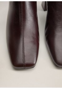 Botas de cuero marrón oscuro hasta el tobillo con punteras cuadradas y detalle de costura central sobre un fondo claro.