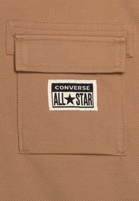 Tessuto marrone con una tasca applicata e una etichetta con il logo "Converse All Star" in bianco e nero cucita sopra. Dettaglio della superficie strutturata visibile.