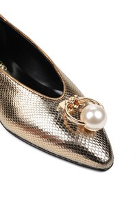 Złote metaliczne buty na wysokim obcasie w szpic z teksturowanym wykończeniem, z dużą perłą przymocowaną złotym pierścieniem na przodzie. Czarny wnętrze.