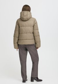 Khaki pehme puffer jope kapuutsiga, millel on tikitud osad. Kantakse hallide pükste ja läikivate mustade saapade peal. Ükslane valge taust.