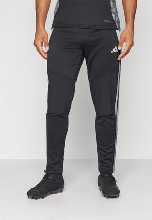 Mężczyzna w czarnych sportowych spodniach Adidas z trzema białymi paskami po bokach i czarnych korkach sportowych, stoi na jednolitym tle.