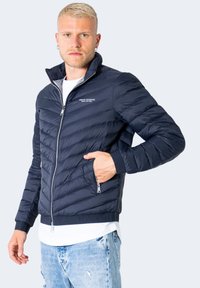 Giacca puffer navy con motivo a chevron, chiusura con zip, colletto alto e tasche laterali, realizzata in tessuto leggero e trapuntato.