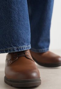 Chaussures en cuir marron avec lacets, présentant une finition lisse et une semelle en caoutchouc robuste, associées à un jean en denim bleu foncé.