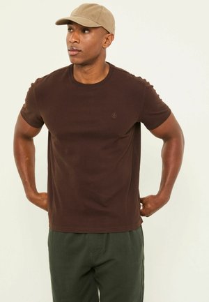 Mężczyzna w beżowej czapce, ciemnobrązowej koszuli z krótkim rękawem i zielonych spodniach, stoi z rękami na biodrach, patrząc na bok.