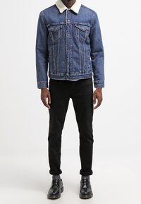 Modrá džínsová bunda s huňatým bílým límcem, s náprsními kapsami a knoflíkovým zapínáním, v kombinaci s černými slim-fit kalhotami a černými botami.