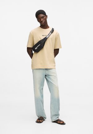 Jonge man die een beige T-shirt, lichtblauwe spijkerbroek, zwarte sandalen en een zwarte schoudertas met witte tekst draagt, staand tegen een witte achtergrond.