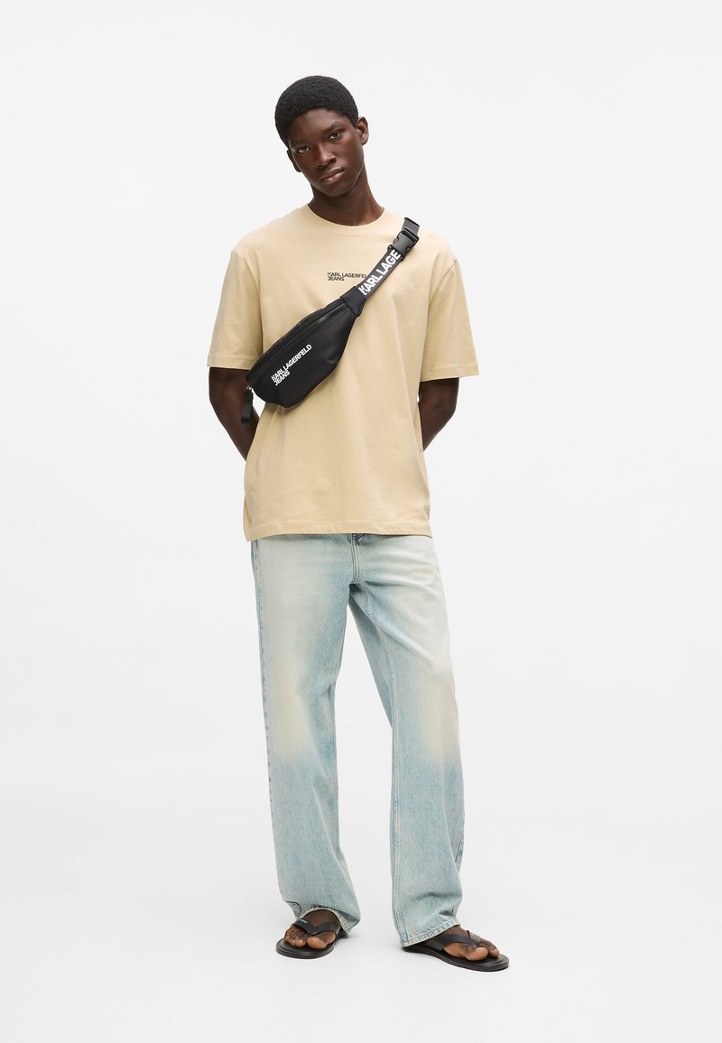 Giovane uomo che indossa una maglietta beige, jeans azzurri, sandali neri e una borsa a tracolla nera con testo bianco, in piedi davanti a uno sfondo bianco.