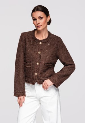 Kobieta ubrana w brązową, teksturowaną kurtkę zapinaną na guziki z złotymi akcentami oraz białe spodnie z wysokim stanem, pozująca z jedną ręką na biodrze.