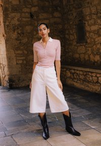 Femme debout contre un mur de pierre, portant un polo rayé rouge et blanc, un pantalon blanc large raccourci et des bottes de cowboy noires.