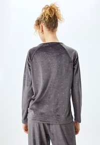 Person med lockigt hår uppsatt i en grå långärmad topp och byxor med små vita prickar, sedd bakifrån.