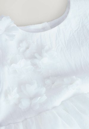 Weißes Kleid mit strukturierter Stoffqualität, rundem Ausschnitt und geschichtetem Tüllrock. Geschmückt mit Stoffblumen in unterschiedlichen Größen.