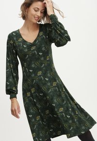 Grün gemustertes Kleid mit langen Ärmeln, V-Ausschnitt, fließendem Rock und strukturiertem Stoff mit gelben und weißen botanischen Designs.