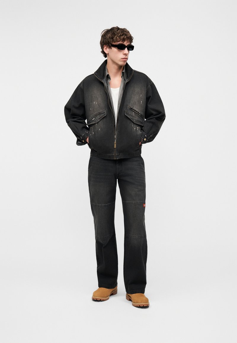 Giovane uomo con giacca e pantaloni di jeans neri sbiaditi, camicia bianca, stivali marroni chiari e occhiali da sole neri, in piedi con le mani in tasca.