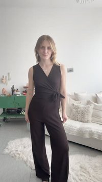 Jeune femme aux cheveux blonds portant une combinaison noire sans manches avec ceinture, debout dans un salon lumineux et moderne avec un canapé blanc.