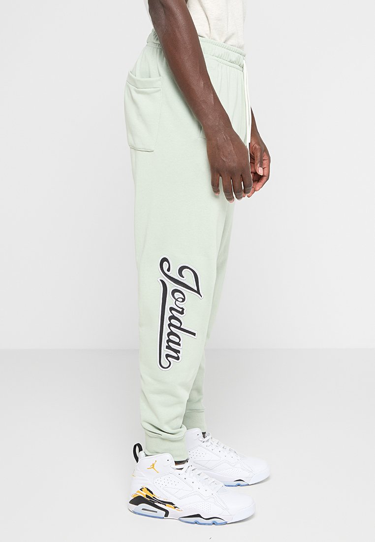 Pantaloni jogger verde chiaro con il logo "Jordan" nero sulla gamba sinistra, dotati di un elastico in vita e tasche laterali, abbinati a sneaker bianche.