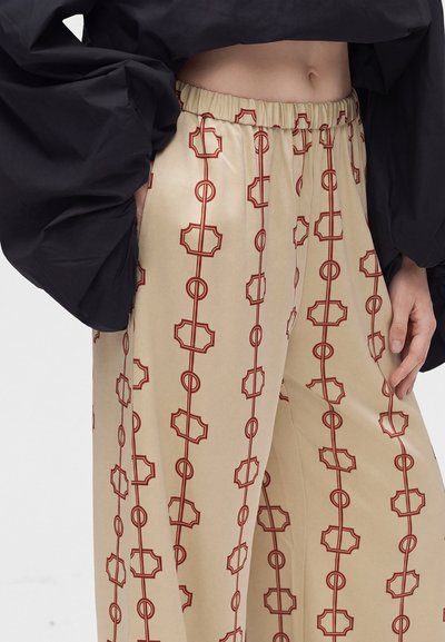 Pantalon beige à jambes larges avec motifs géométriques rouges en forme de chaîne et taille élastique, porté avec un haut noir à manches longues découvrant le ventre.