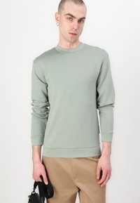 Sweatshirt verde claro com mangas longas, punhos canelados, gola arredondada e uma textura suave. Combinado com calças bege e um acessório na mão.