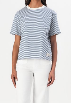 Person iført en kortærmet, marineblå og hvidstribet skjorte med hvid krave og hvide bukser, stående mod en ensfarvet baggrund.