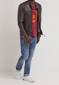 Brun læderjakke med lynlåslommer, båret over en rød T-shirt med et gult emblem. Blå jeans og hvide sneakers fuldender outfittet.