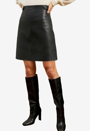 Mini jupe en cuir noir avec une texture lisse, dotée d'une couture sur le devant et d'une quincaillerie décorative à la taille. Associée à des bottes noires jusqu'aux genoux.