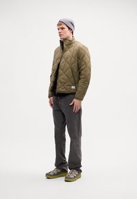 Joven vestido con una chaqueta de oliva acolchada, un gorro gris, jeans gris oscuro y zapatillas con detalles verdes, de pie frente a un fondo blanco liso.