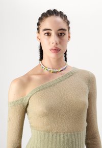 Pleteni, pulover s otvorenim ramenima u mekoj bež boji s teksturiranim rebrastim dizajnom, uparen s necklace od višebojnih perli koje imaju osmijehe.